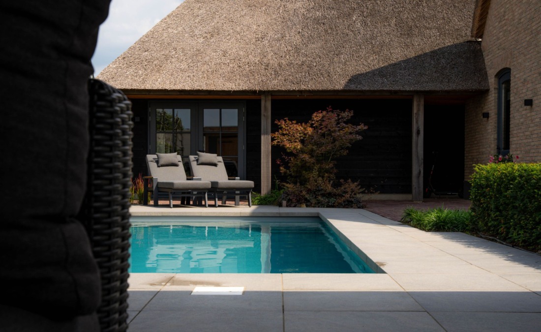 Langwerpig HDPE zwembad met helder blauw water bij luxe villa met rieten kap en twee grijze ligbedden.
