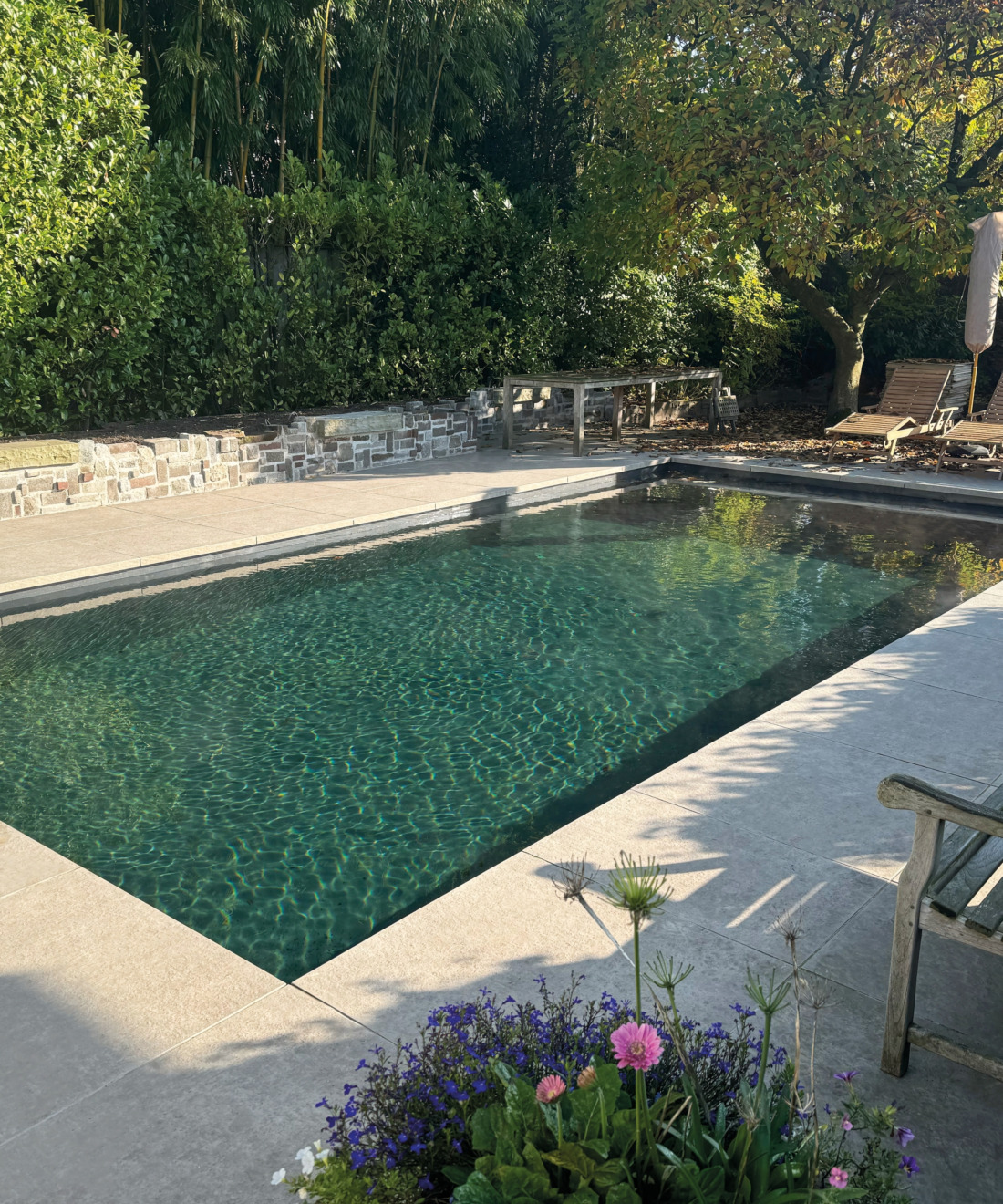 Buitenbad met een soort van groenig kleurend water, omringd door grote tuintegels en houten ligstoelen