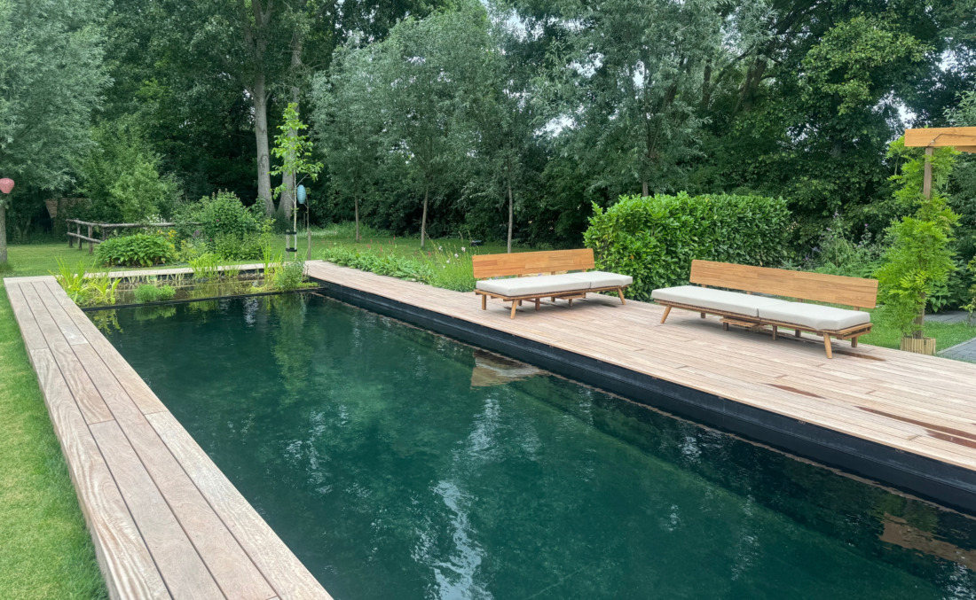 Lange zijde van een zwart HDPE zwembad in zwemvijver-stijl, voorzien van een breed houten vlonderterras met loungebanken.