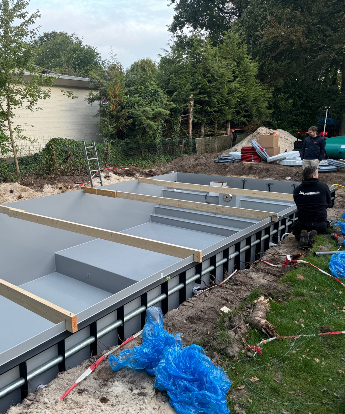 Installatie van een grijze zwembadkuip in de tuin, verstevigd met houten dwarsbalken tijdens het aanvullen van de grond.