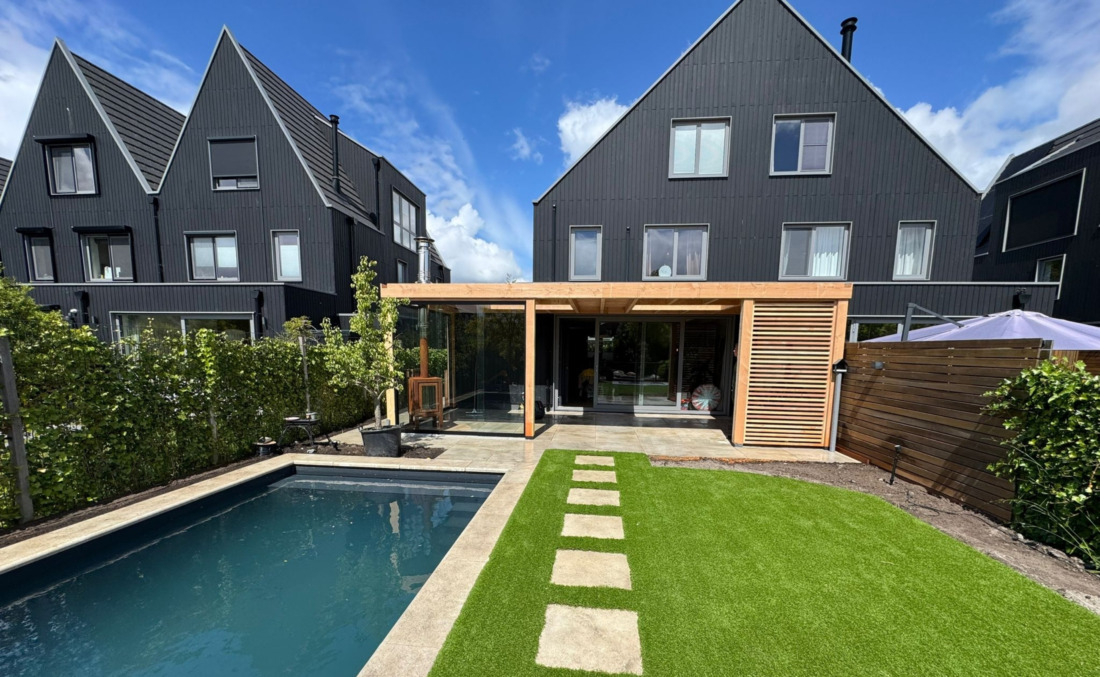 Strak zwart HDPE zwembad van JDL in een moderne tuin bij zwarte nieuwbouwwoningen, grenzend aan een luxe houten tuinkamer met glazen wanden.